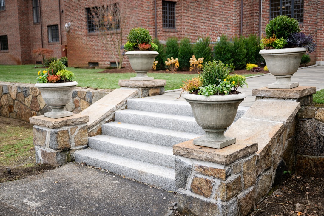 Stone steps with four planters featuring greenery lead to a brick building, surrounded by a well-maintained garden.