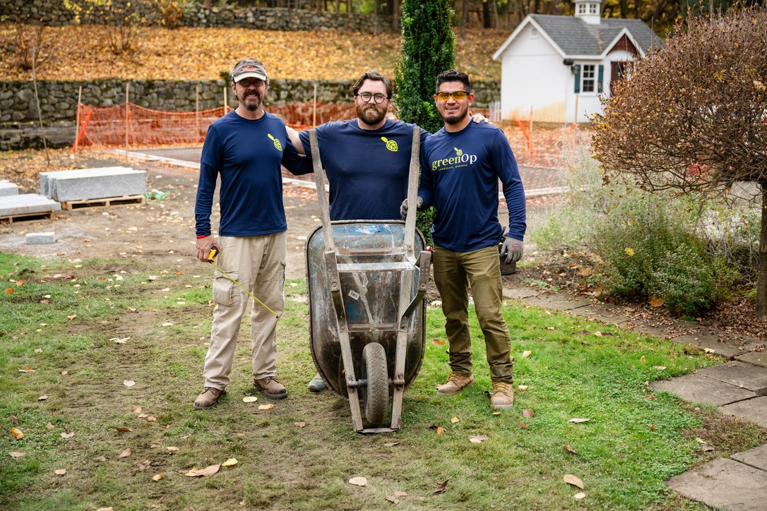 Three people in blue shirts stand with a wheelbarrow on grass, next to a white shed. Orange safety fencing and stone wall in background.