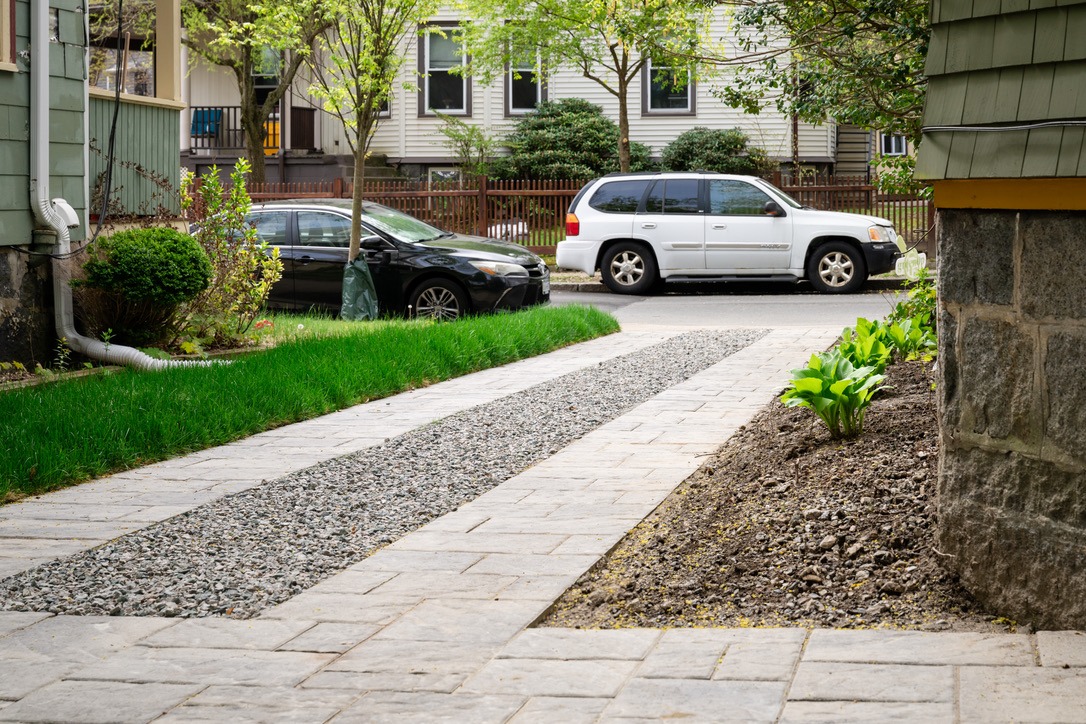 A gravel driveway bordered by plants leads to a residential street with parked cars, trees, and houses in the background.