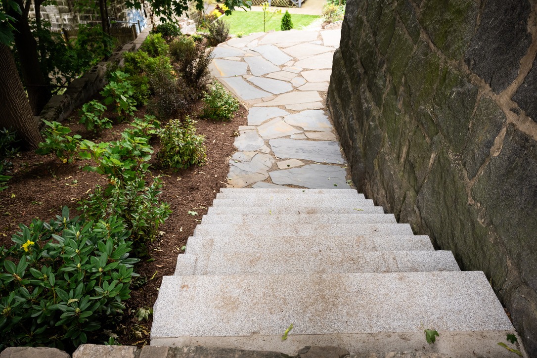 Stone steps descend between lush greenery and a textured stone wall, leading to a paved path surrounded by landscaped garden.