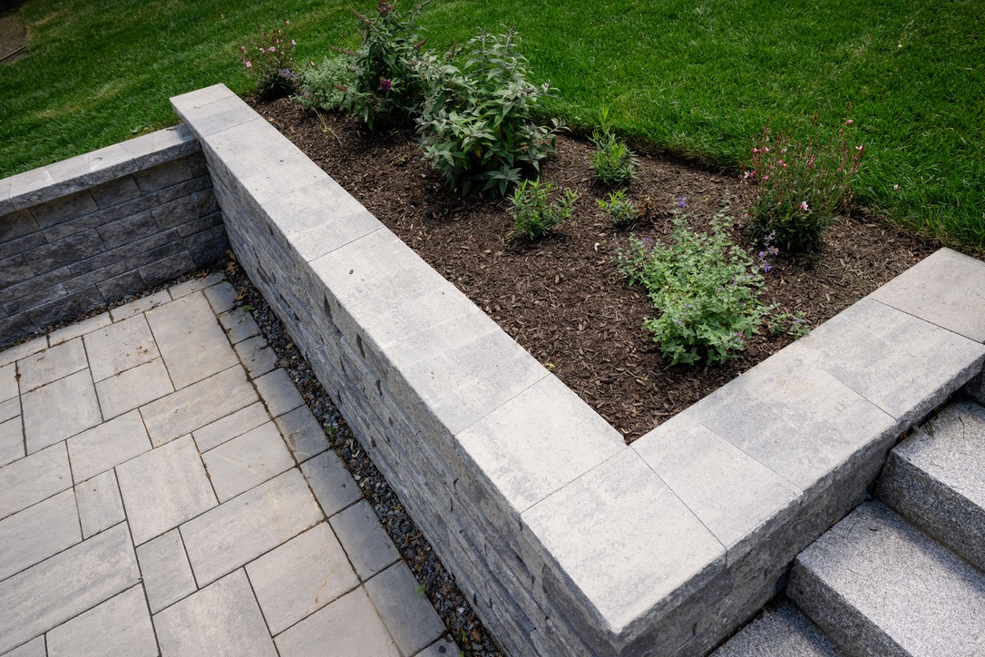 A neatly arranged garden bed with small plants, surrounded by a low stone wall, bordered by neatly laid stone patio and green grass.