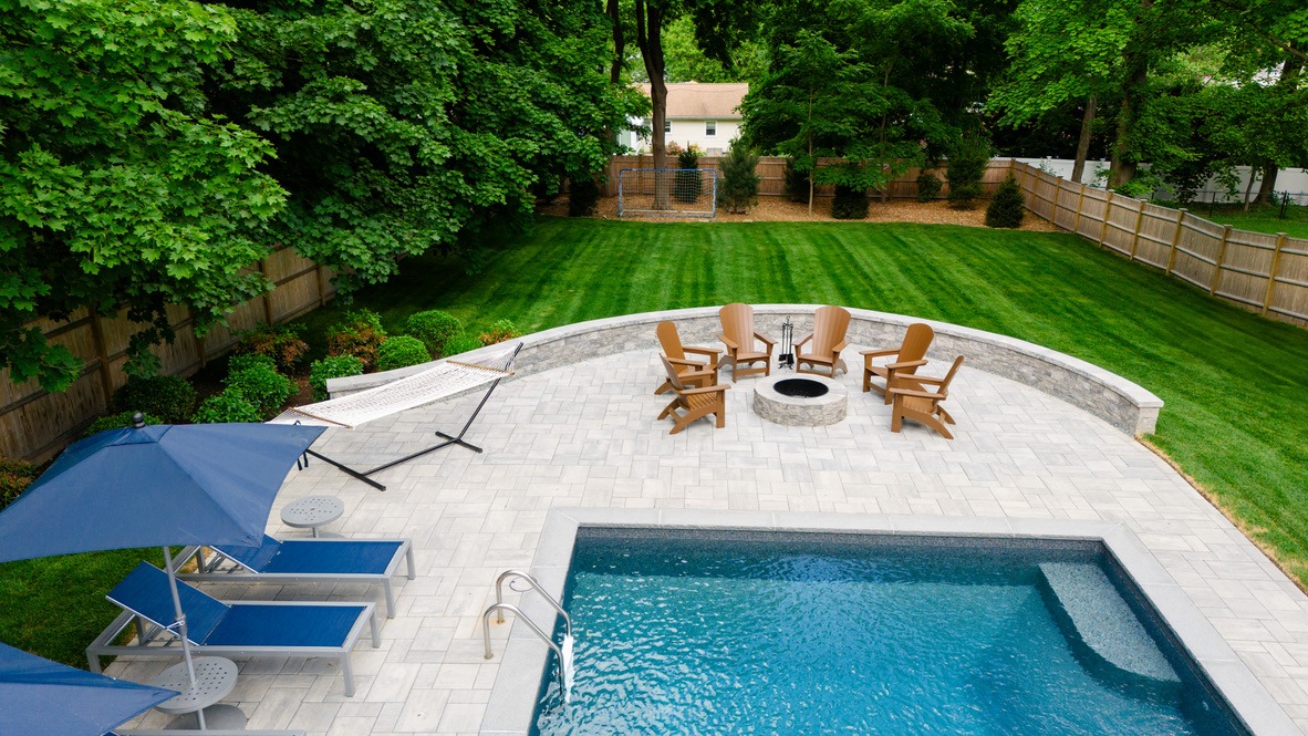 Backyard with a swimming pool, patio, lounge chairs, umbrellas, hammock, fire pit, circular seating, and lush green lawn surrounded by wooden fencing.