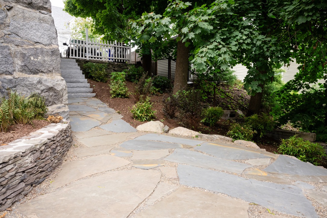 Stone path and stairs leading to a white fence, surrounded by greenery and trees. Peaceful natural setting with a person walking.