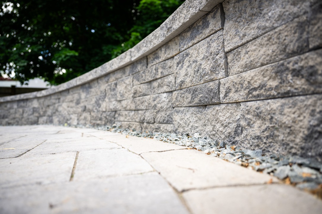 Curved stone wall with textured surface, adjacent to a pathway lined with gravel. Trees in the background add greenery.