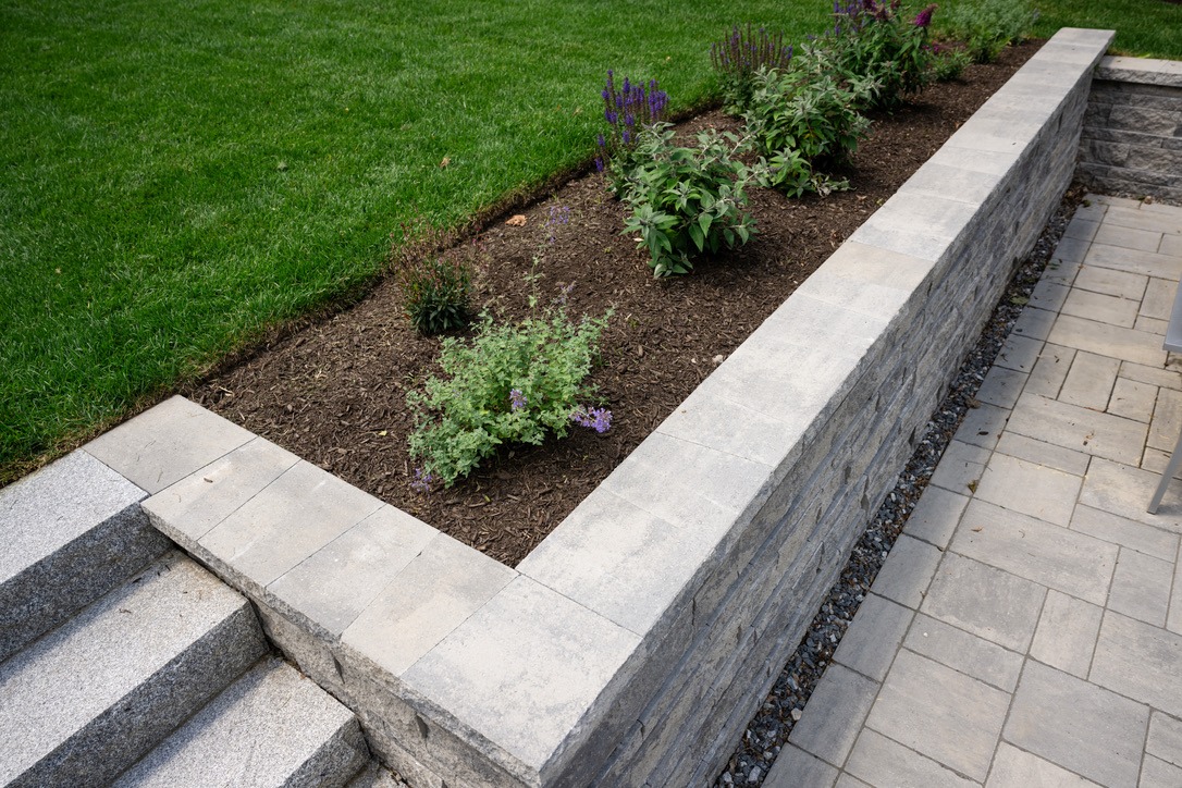 A neatly trimmed garden with various plants in a raised stone bed, adjacent to stone steps and a paved patio area.