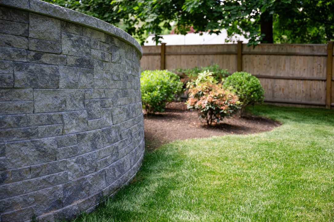 Stone wall curves along a well-maintained garden with shrubs and a wooden fence, surrounded by lush green grass and trees.