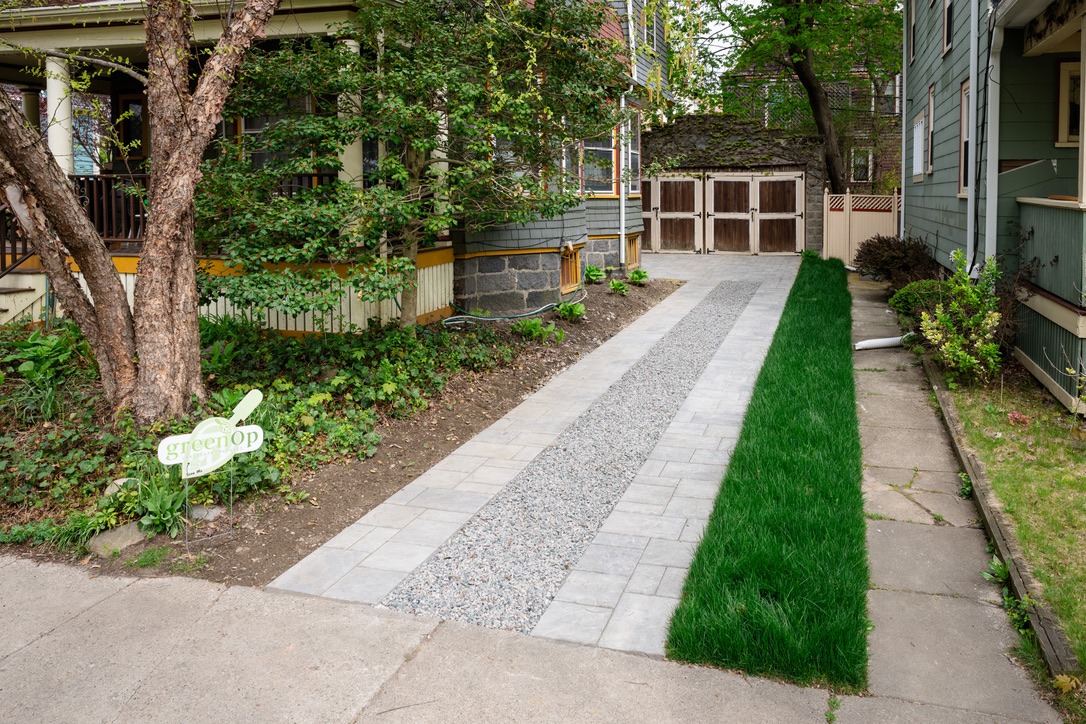 Tree-lined path with gravel and grass, leading to a wooden garage. GreenOp Lawn sign on the left side, between two houses.