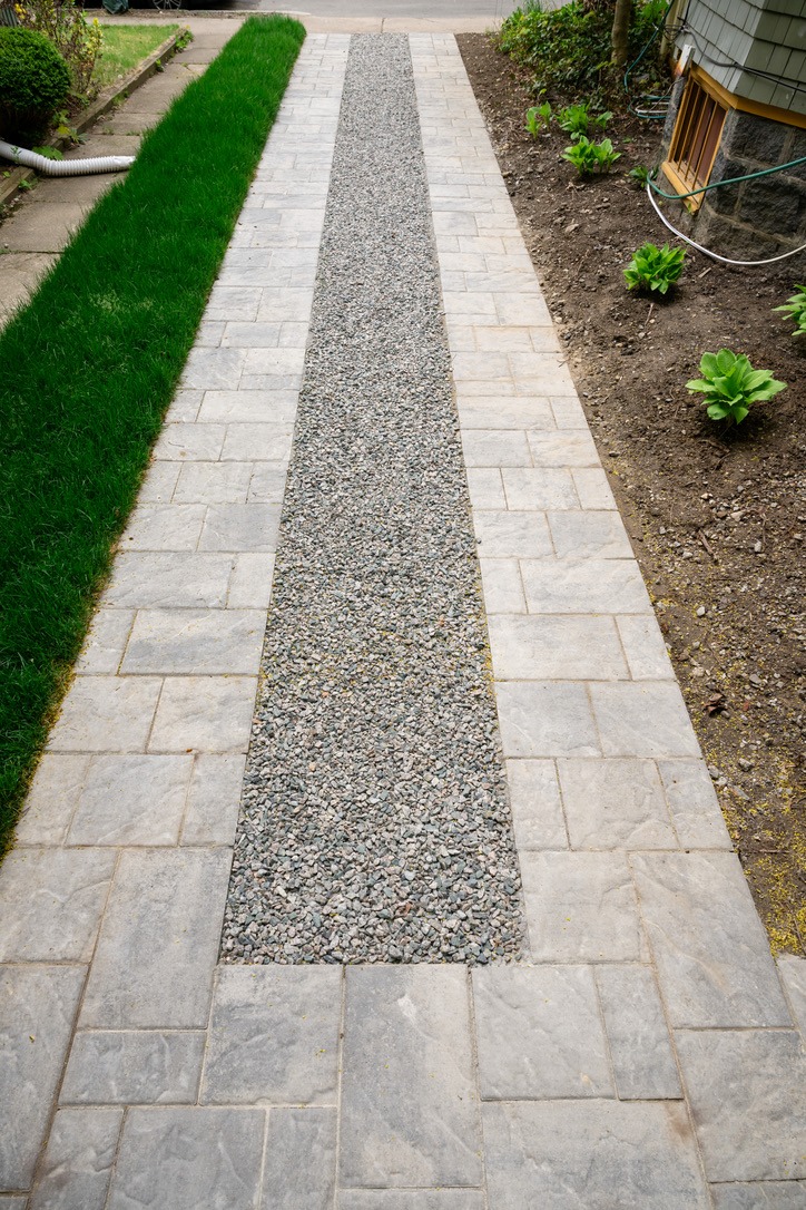 A gravel and stone walkway flanked by green grass and soil, with some small plants, leading to a residential building.