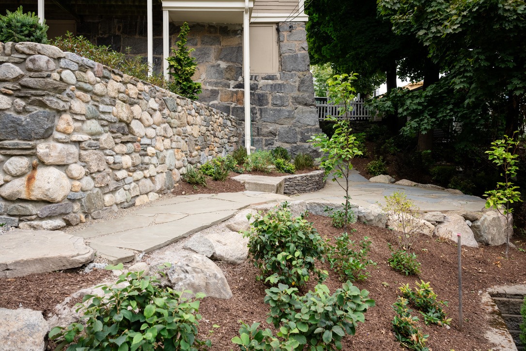 A stone pathway leads through a landscaped garden with shrubs and trees beside a rustic stone wall under a house's porch.