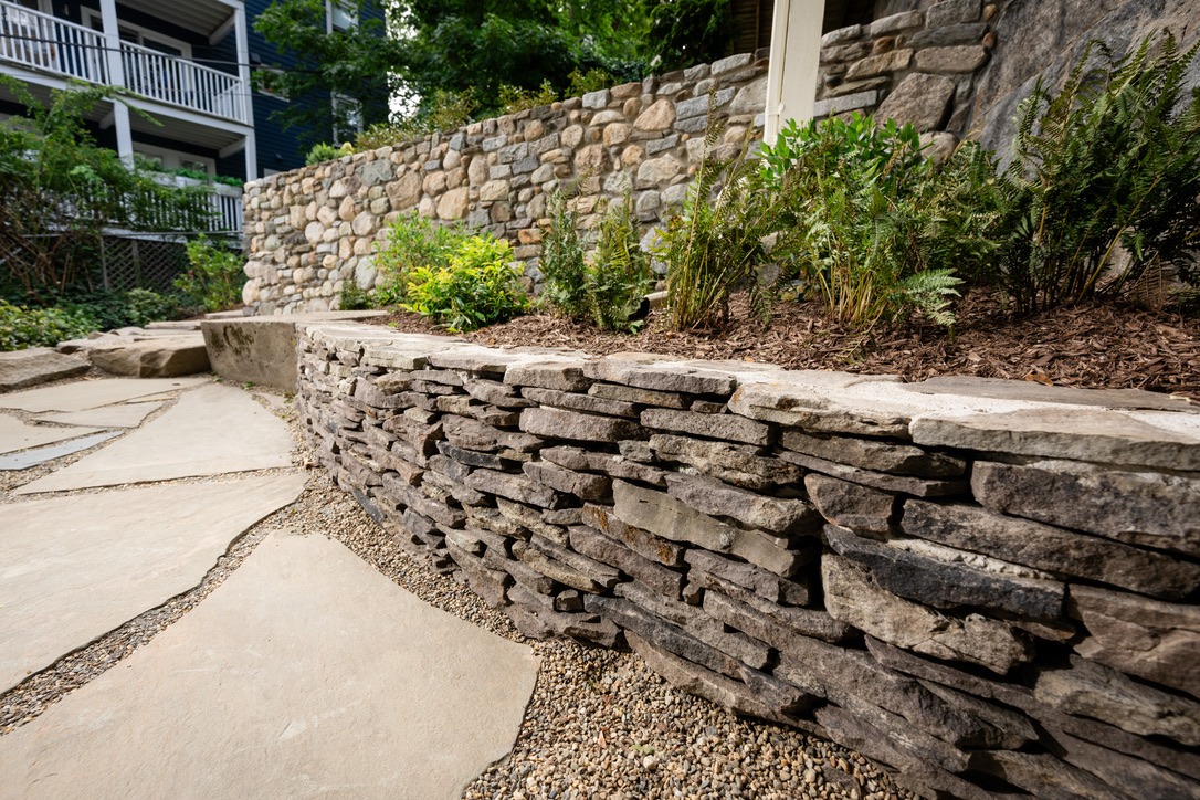 Stone retaining wall with plants, mulched garden bed, and gravel path in a landscaped area. Residential building with balconies in background.