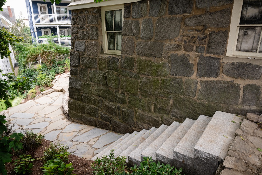 Stone steps descend beside a rustic stone building with plants. Another house and garden are visible in the background.