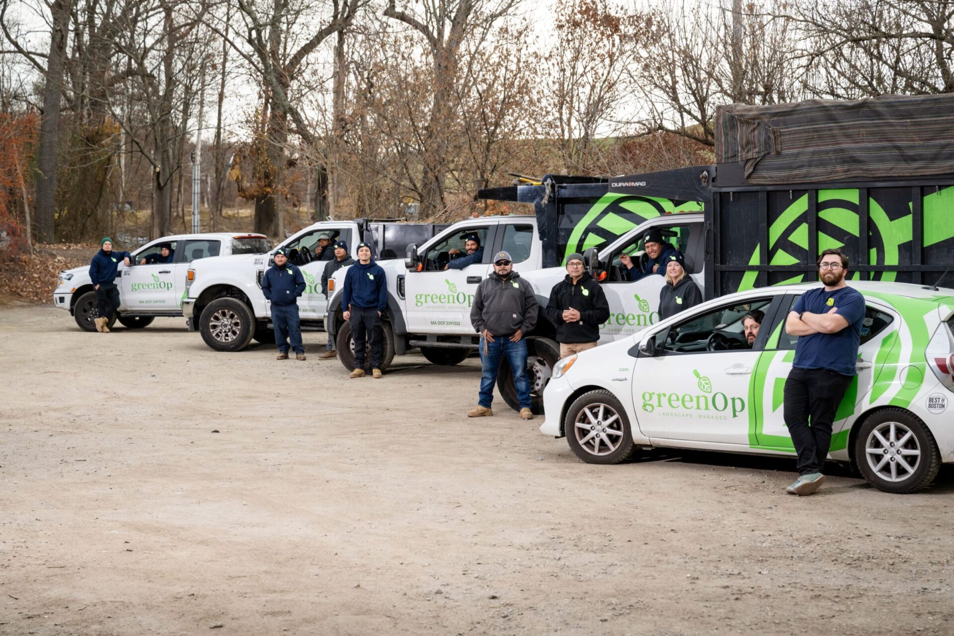 A group of people stands beside vehicles displaying the "greenOp" logo, with a wooded background and overcast sky.