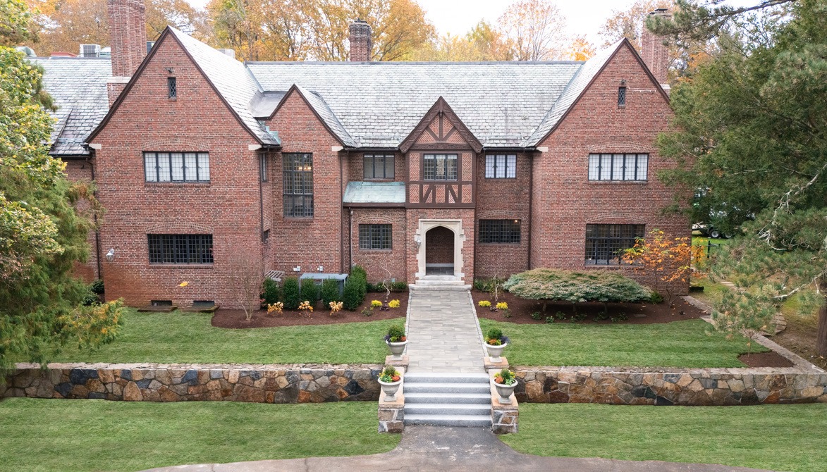 A large brick house with Tudor-style architecture, surrounded by well-maintained gardens and trees, featuring stone steps leading to the entrance.