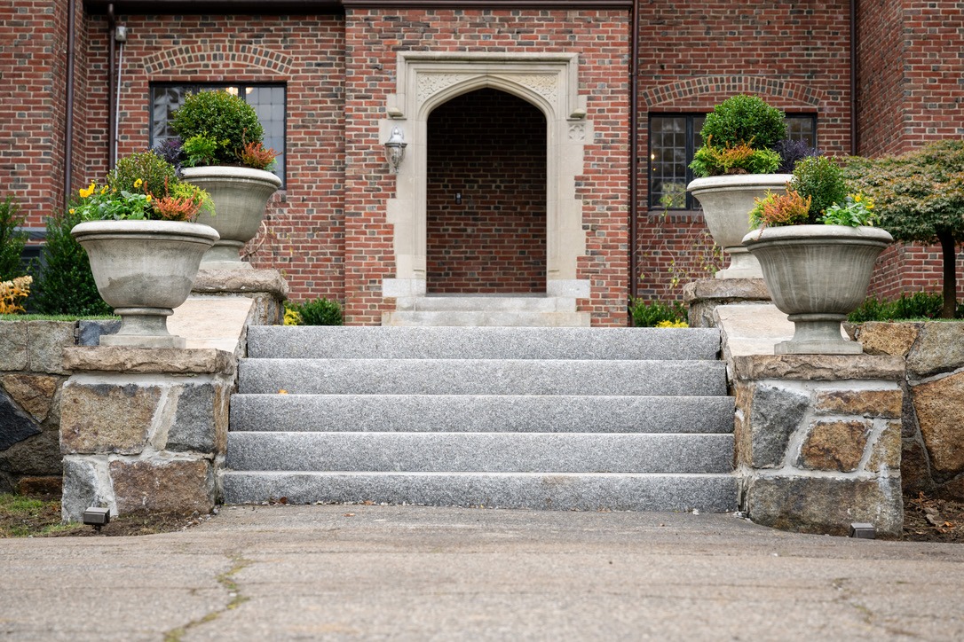 Stone steps lead to a brick building entrance, surrounded by large potted plants and decorative stonework. No recognizable landmarks or historical buildings.