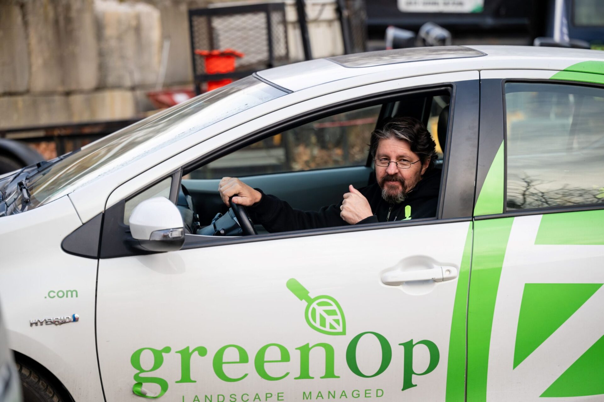 A person inside a white car with greenOp logo, giving a thumbs up, parked near a concrete structure with a red bin nearby.