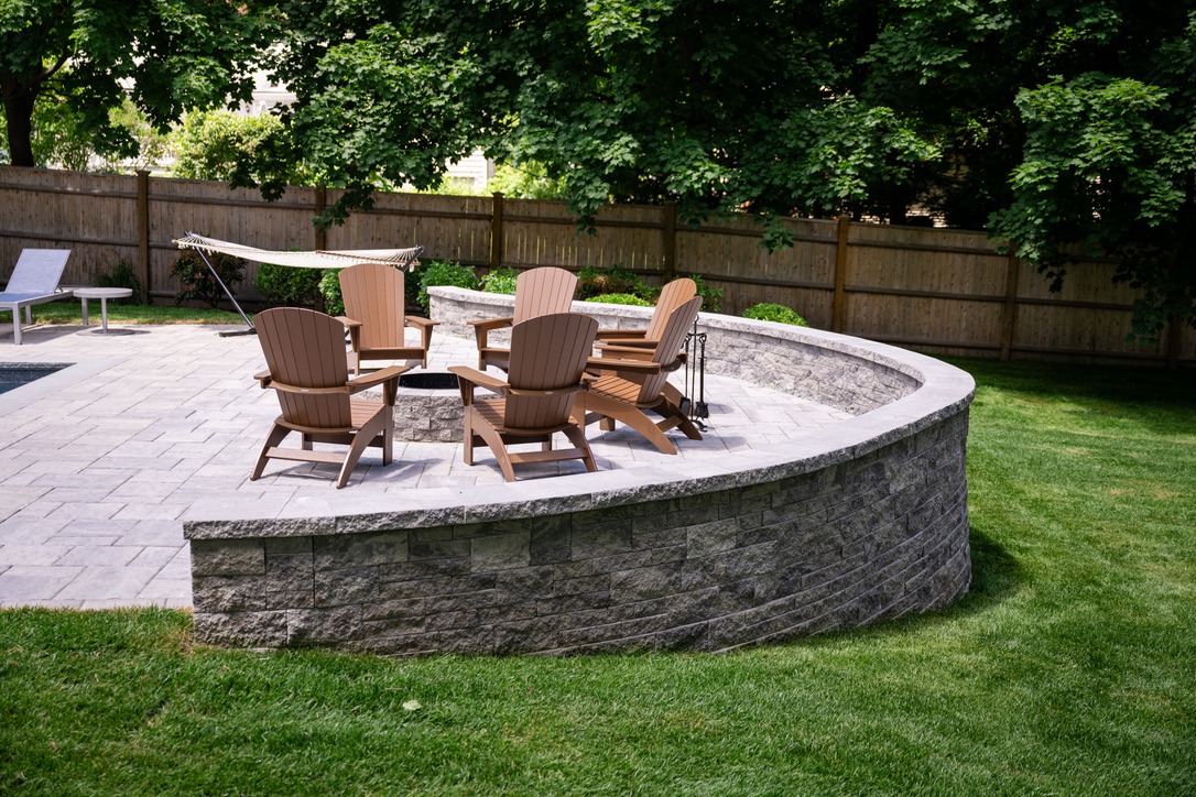 A stone patio features Adirondack chairs around a fire pit, surrounded by trees and a hammock, enclosed by a wooden fence.