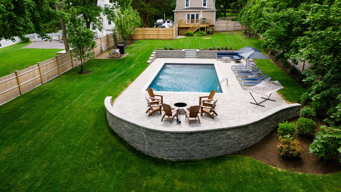 A fenced backyard features a rectangular pool, surrounded by lounge chairs, a hammock, and seating area, with adjacent house and lush greenery.