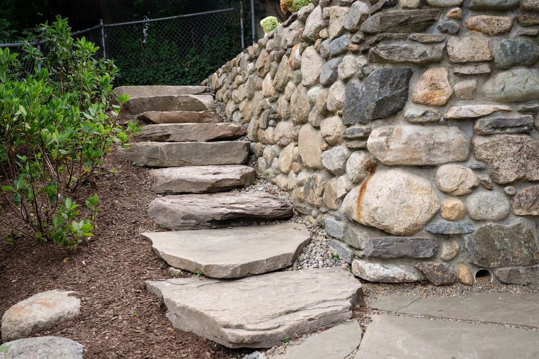 Stone steps lead up beside a rustic rock wall, accompanied by lush greenery and fenced background, creating a natural, serene garden path.