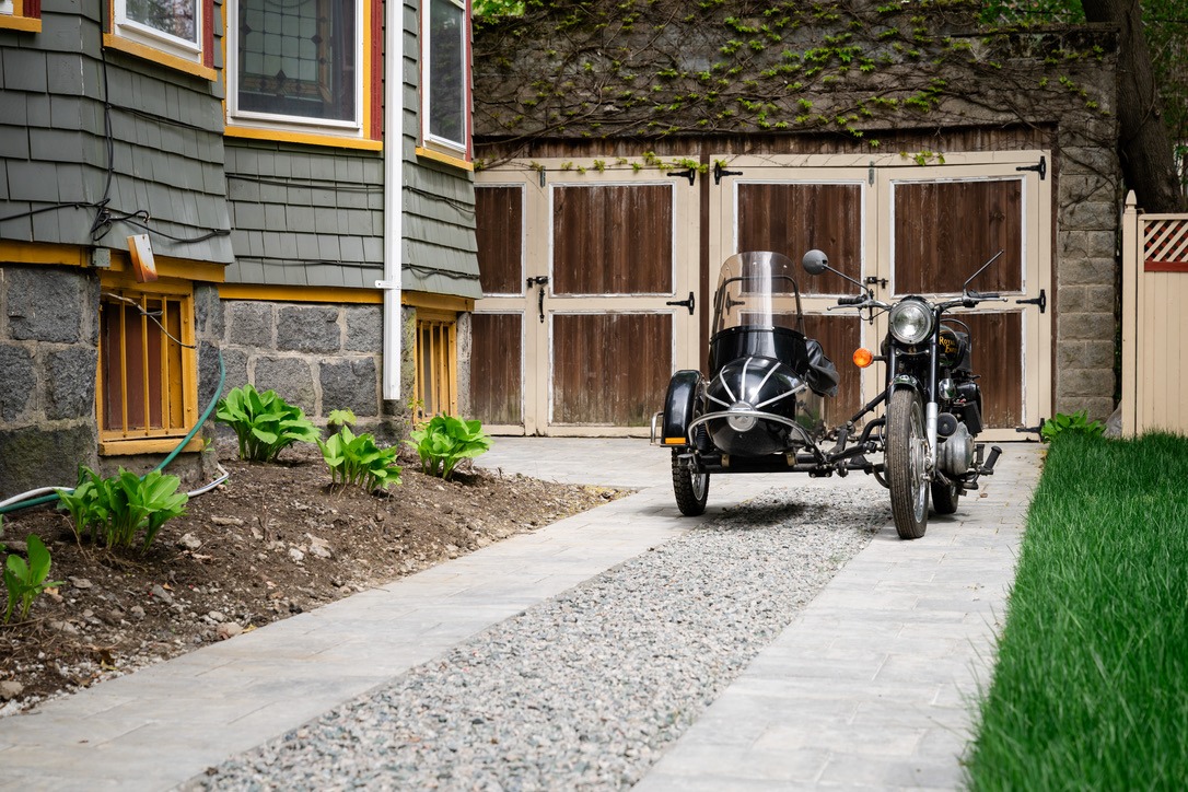 A classic motorcycle with a sidecar is parked on a stone pathway beside a charming house with wooden double garage doors.