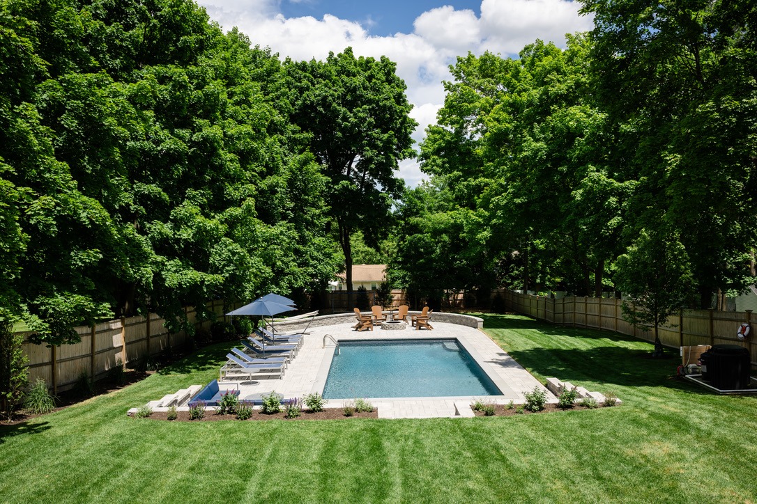 A backyard pool surrounded by lush trees, sun loungers, and a patio area with chairs. Fenced, with neatly trimmed grass and plants.