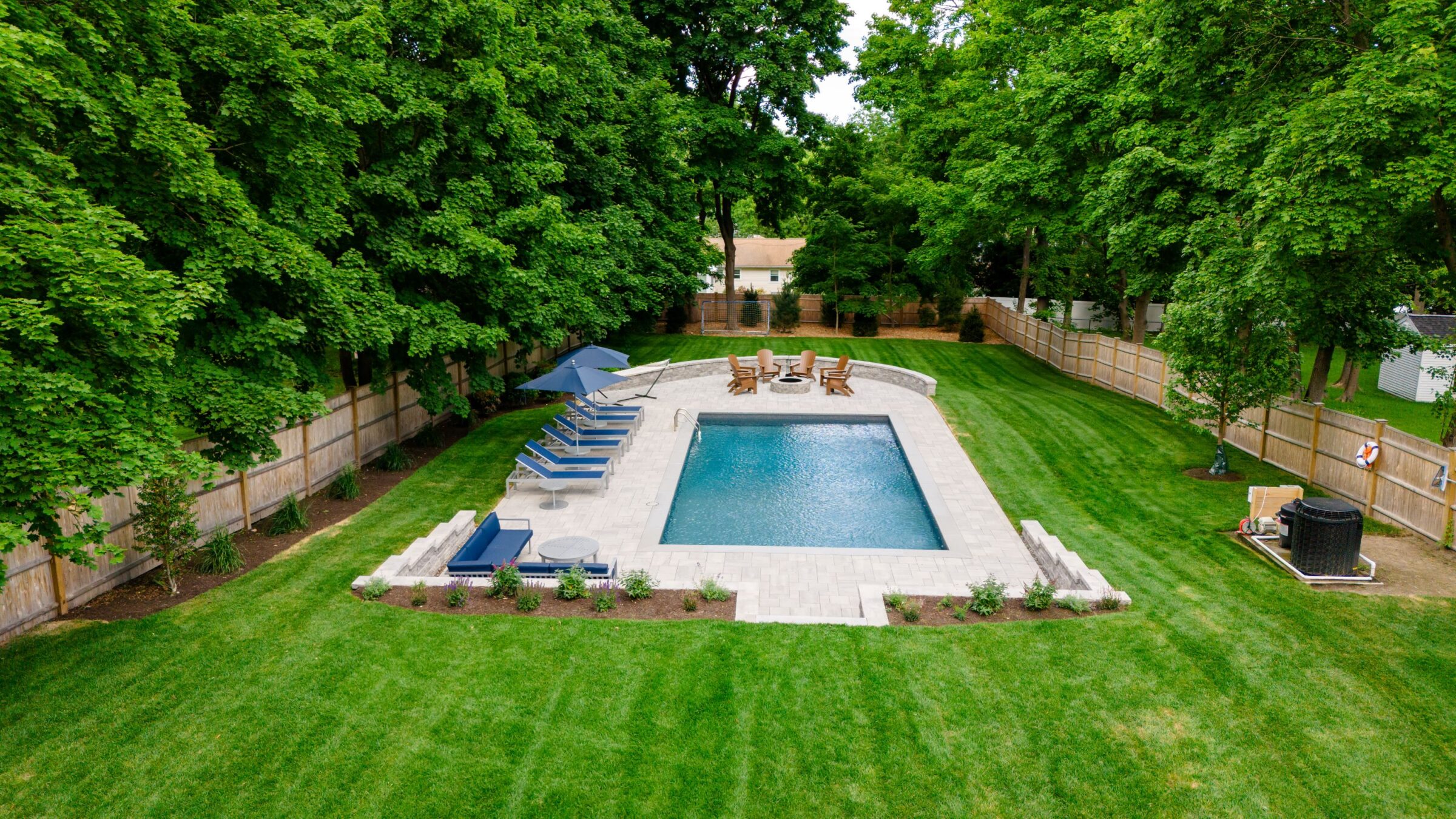 Aerial view of backyard swimming pool surrounded by lounge chairs, lush green lawn, and trees. Wooden fencing encloses the tranquil outdoor space.