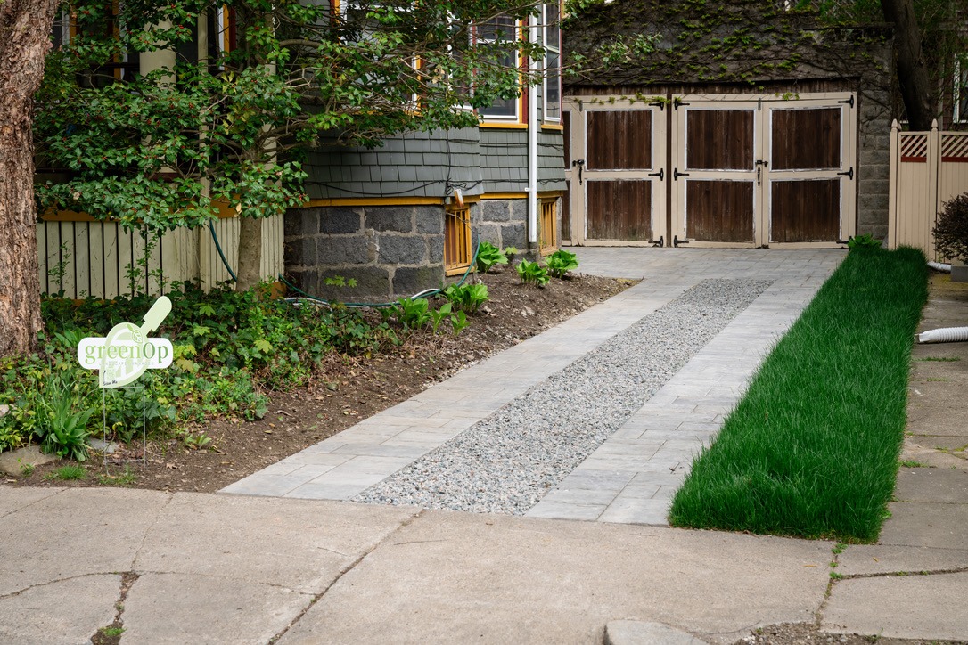 A driveway with stone and gravel path, bordered by green grass and shrubs, leads to a wooden garage. Sign reads 