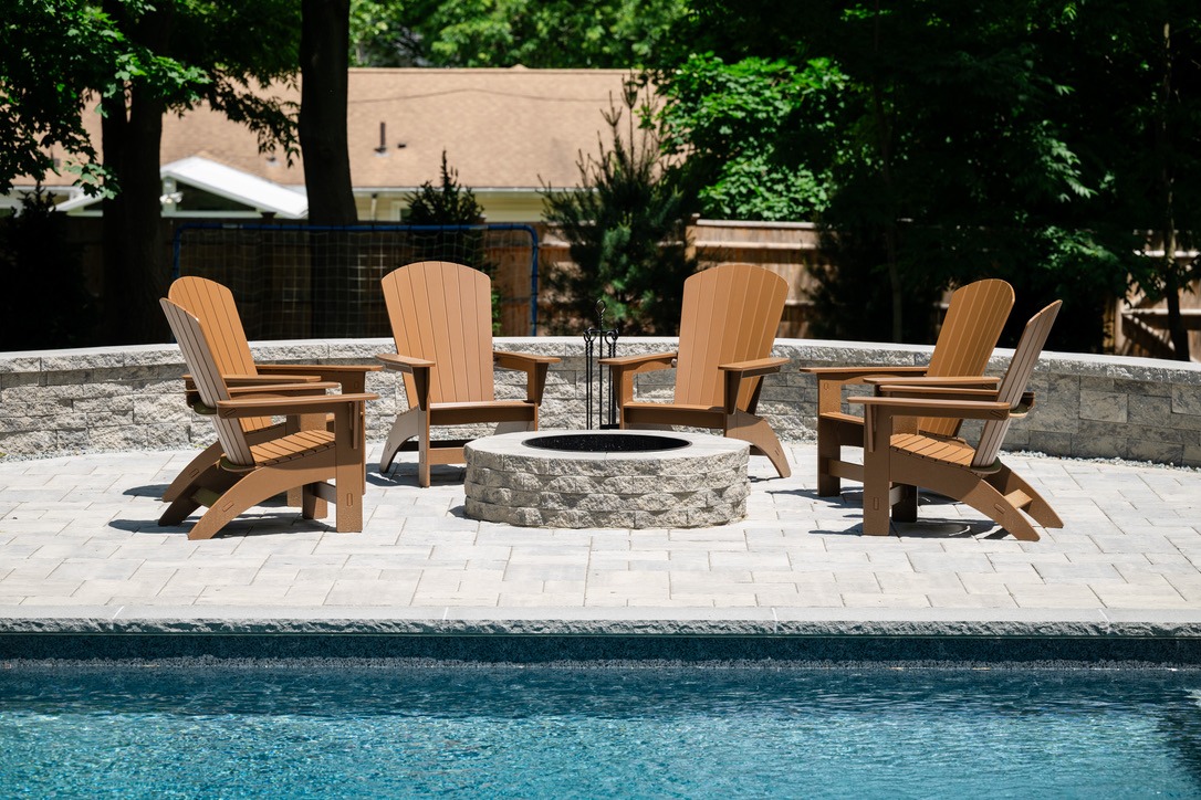 Five wooden chairs encircle a stone fire pit on a patio by a pool, surrounded by trees and a fence in the background.