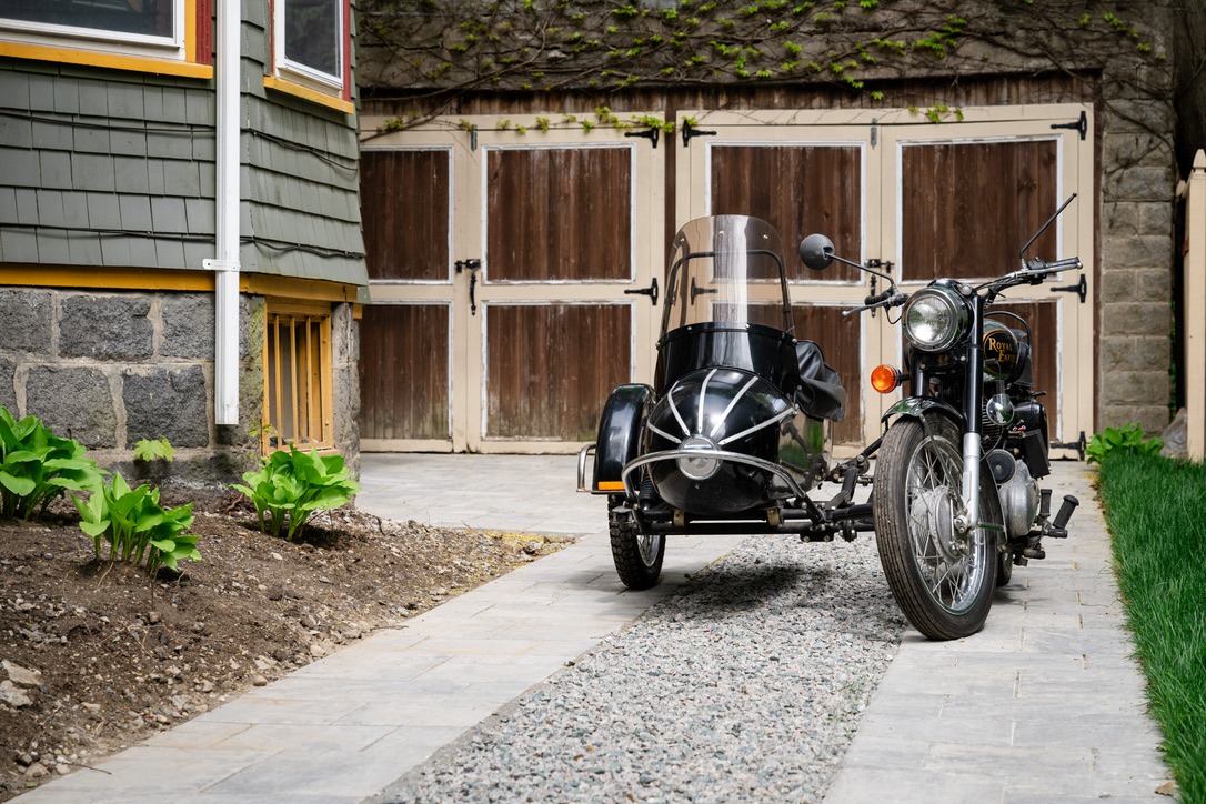 A vintage motorcycle with sidecar is parked on a stone driveway beside a house with green siding and wooden garage doors.