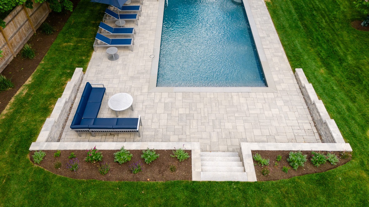 Modern backyard with a rectangular pool, blue seating, lounge chairs, and landscaped garden. Surrounded by grass and a wooden fence.