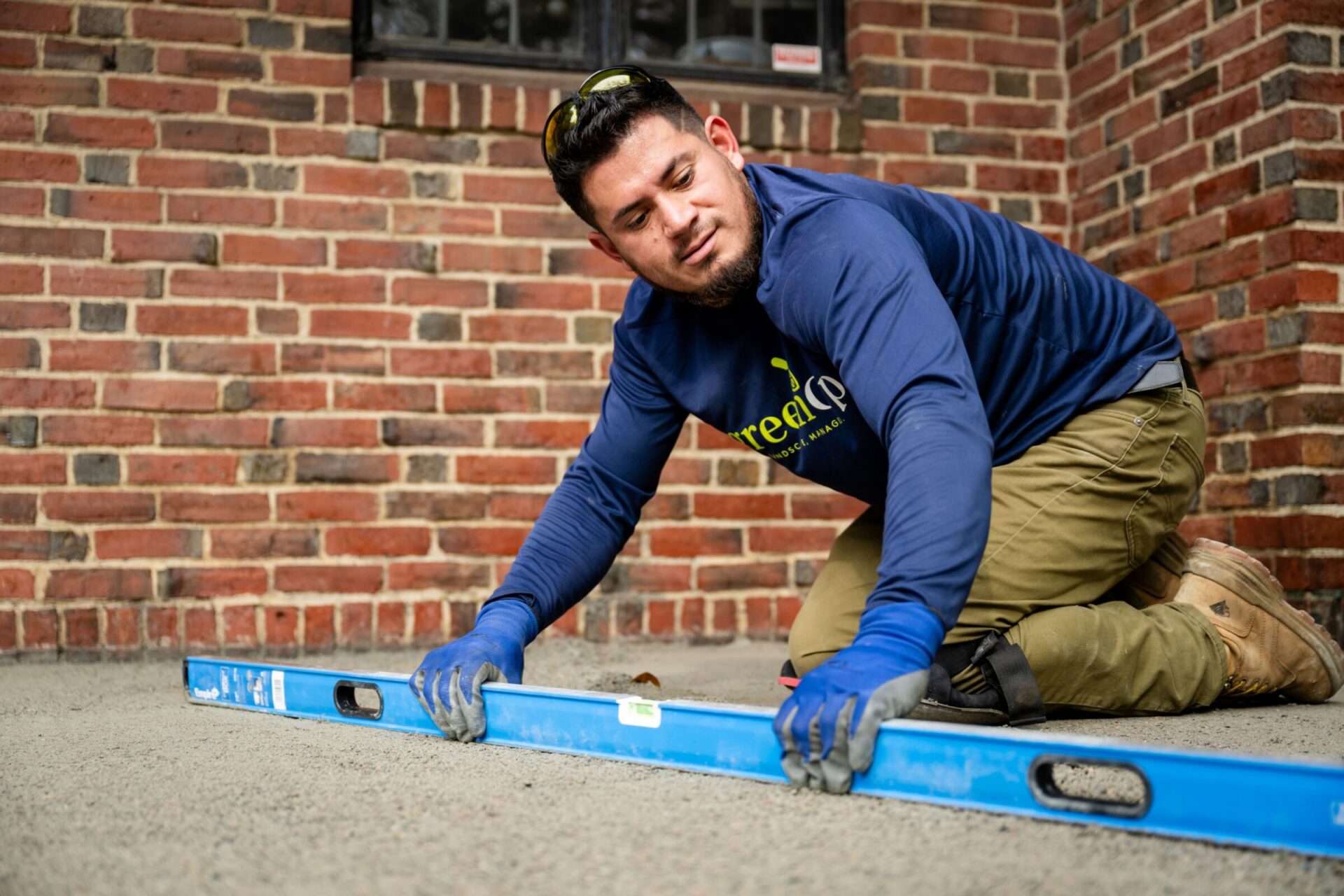 A person in gloves and boots levels a surface with a blue tool against a brick wall background.