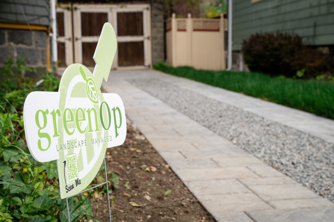 A green landscape company's sign on a yard beside a paved path, leading to a wooden gate and fence.