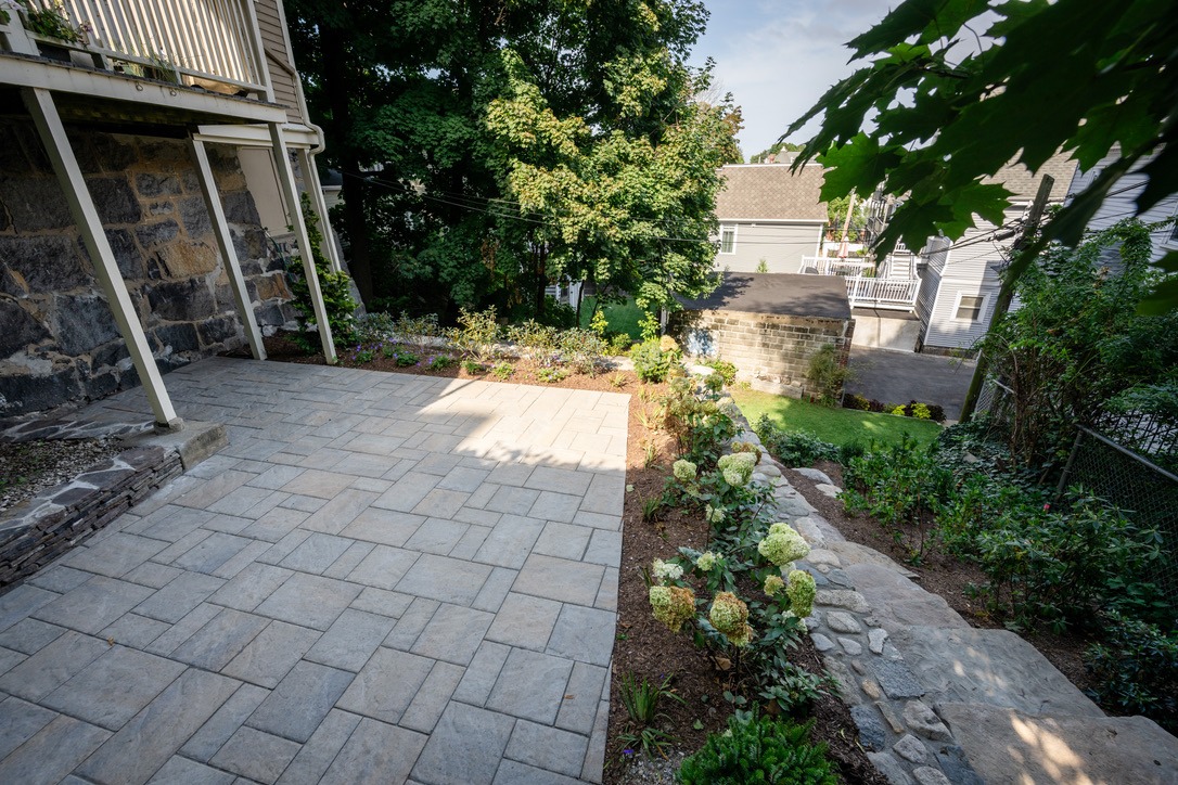 A stone patio beside a building features landscaped flower beds and leads to a garden area with trees and neighboring houses.