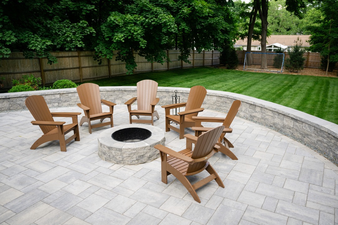 A backyard patio features six wooden chairs around a stone fire pit, surrounded by lush grass, trees, and a wooden fence.