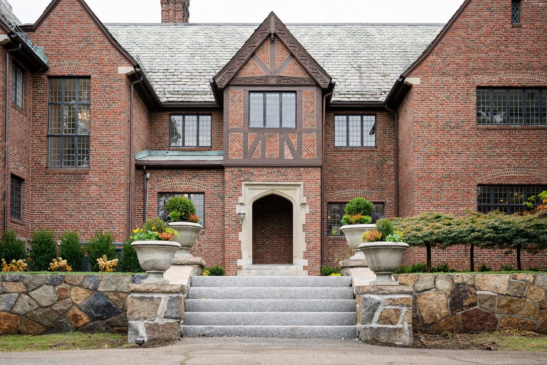 A Tudor-style brick mansion with detailed woodwork, stone steps, and decorative planters on either side, leading to an archway entrance.