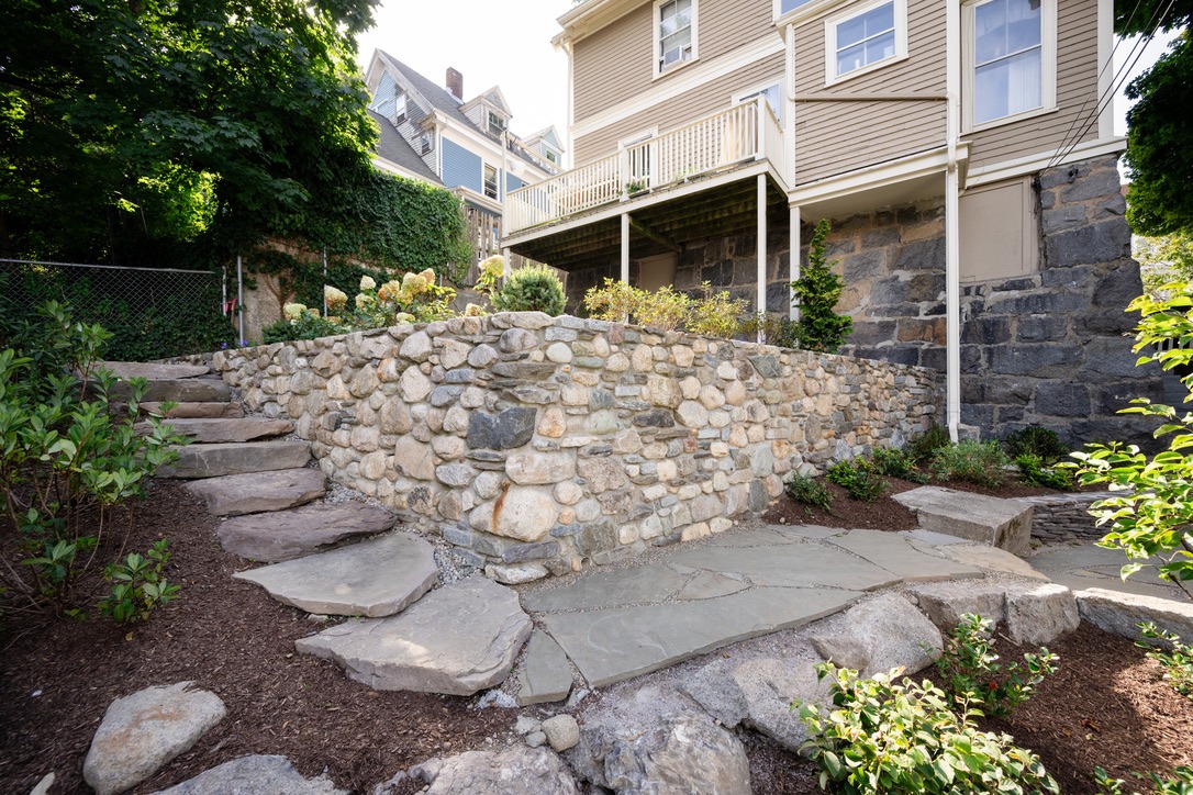 A stone walkway with steps leads to a house with a raised garden, surrounded by lush greenery and a stone retaining wall.