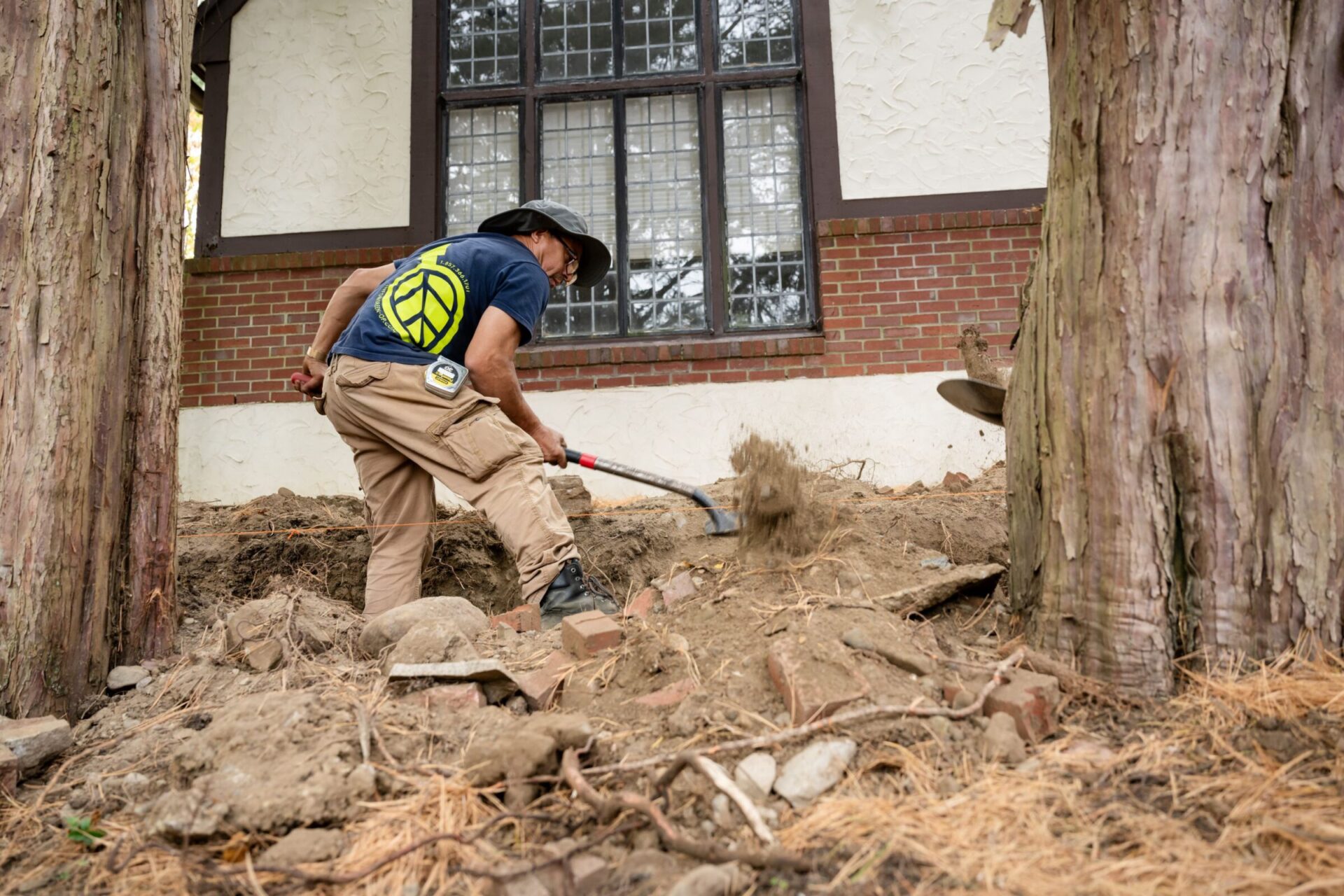 A person is digging near a historic brick building, wearing outdoor work clothing. Large trees and exposed roots surround the excavation site.