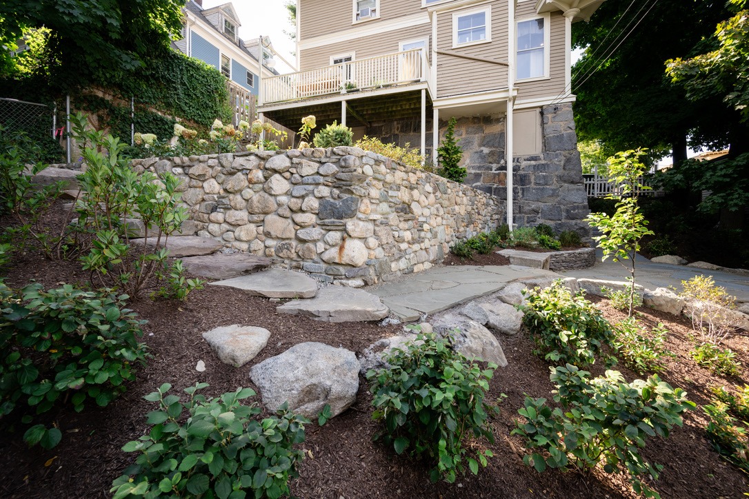 A stone retaining wall surrounds a landscaped garden beside a wooden house, with lush greenery and a tidy pathway leading upward.