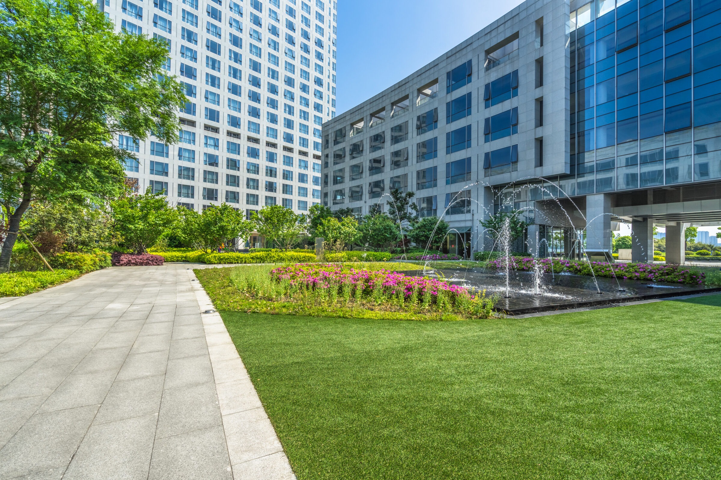 Modern office buildings with glass facades surround a tranquil garden featuring a pathway, colorful flowers, green lawn, and a central fountain.